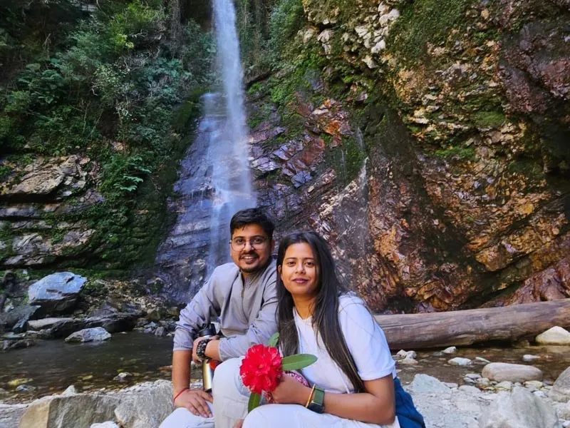Couple sitting together on a fallen log in front of a tall cascading mountain waterfall in Jibhi