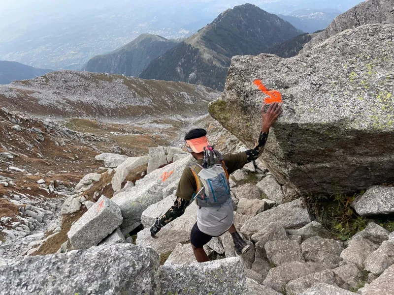 Hiker scrambling over rocky boulder ridge near Indrahar Pass trail marker flag