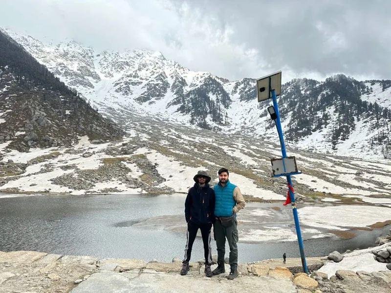 Two trekkers standing by a mountain trail marker against snow-capped peaks at Kareri Lake