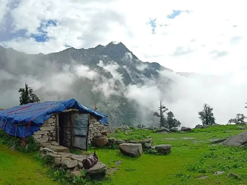 Rustic stone shepherd hut in a green alpine meadow surrounded by mist and clouds