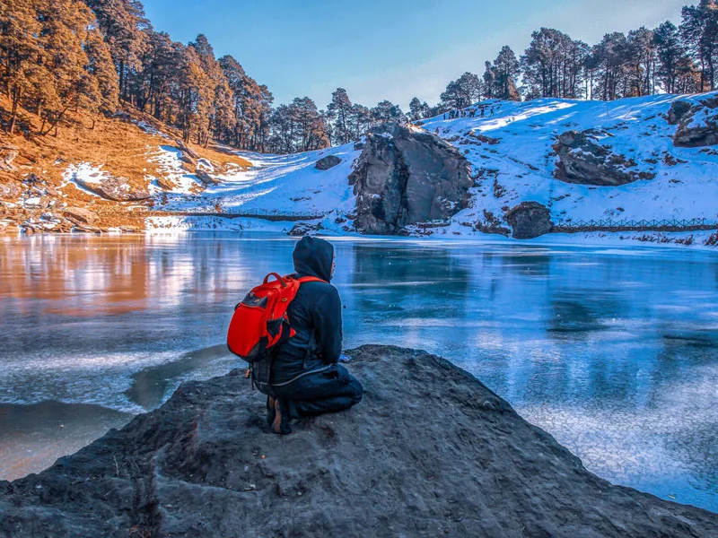 Trekker sitting on a rocky ridge overlooking partially frozen Serolsar Lake and snow-covered trees