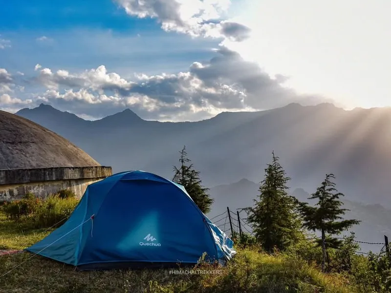 Blue alpine dome tent pitched on a scenic grassy ridge overlooking misty valleys and sunburst rays